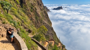 Hiker photographing a spectacular mountain trail above a sea of clouds in Cape Verde, surrounded by dramatic cliffs and volcanic landscapes.