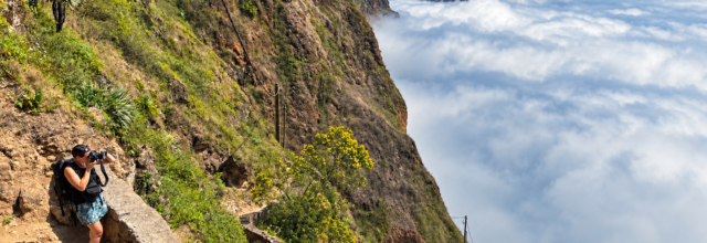 Hiker photographing a spectacular mountain trail above a sea of clouds in Cape Verde, surrounded by dramatic cliffs and volcanic landscapes.